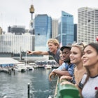 Excited multi-ethnic friends looking away against buildings. Happy males and females are standing on bridge over river. They are traveling together in city.