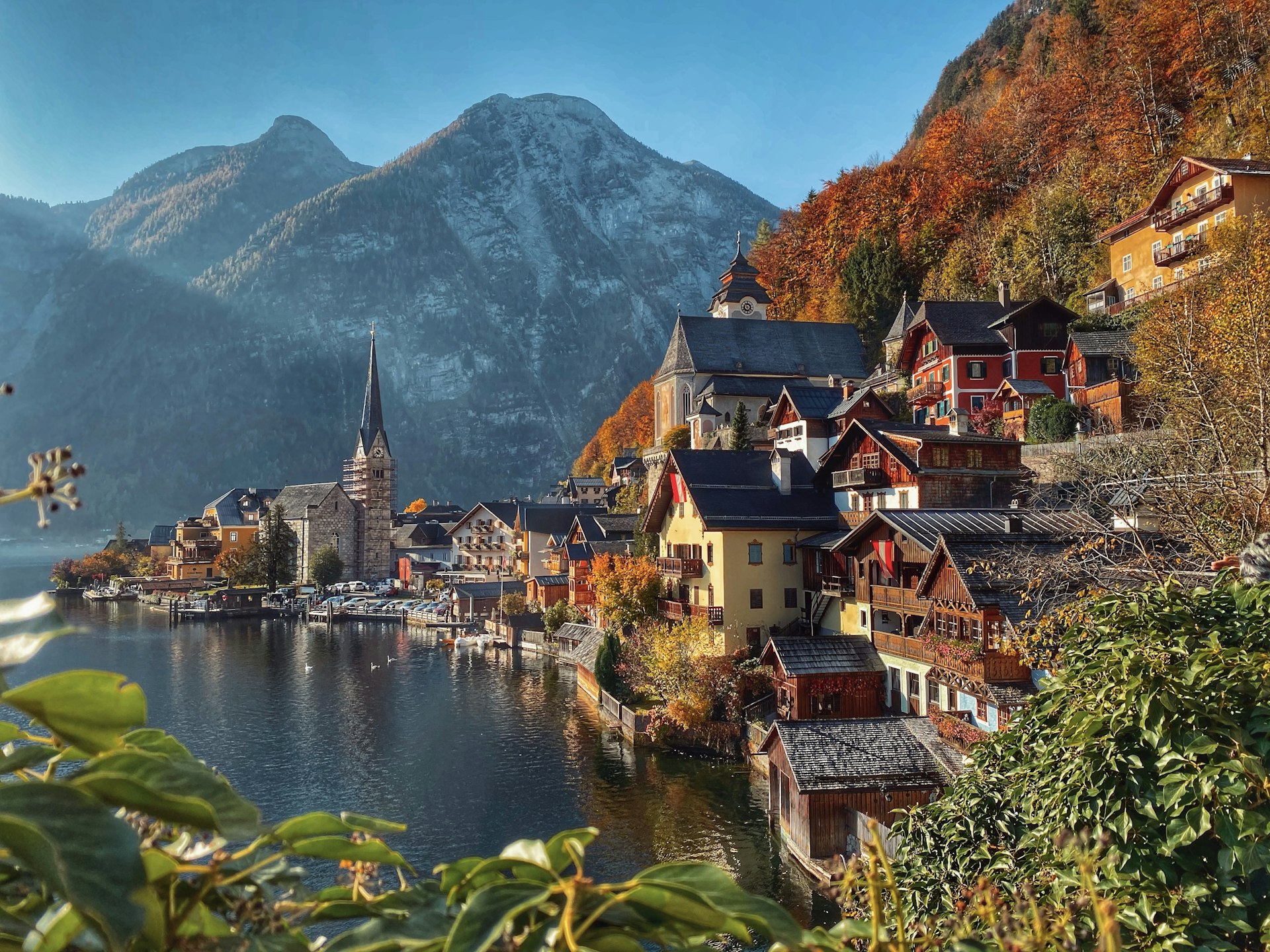 Historic Hallstat sits on the edge of a flat, reflective lake, with mountains in the background. The leaves are bright orange and red.